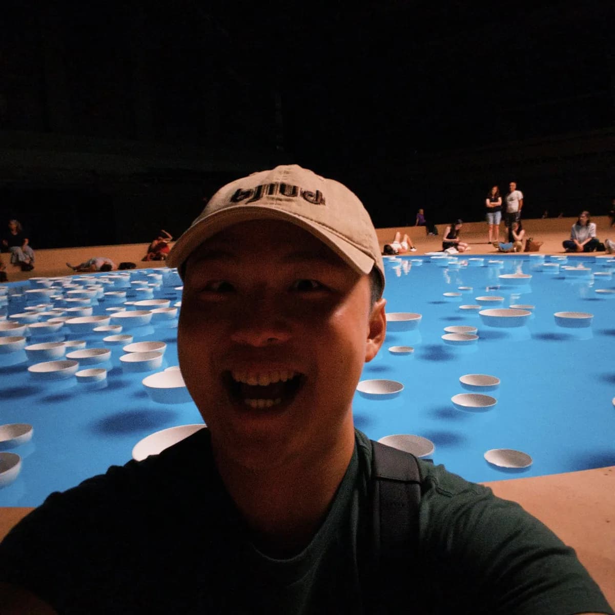 Chi Kang Pai grinning at the camera in front of a large installation: a blue floor covered in white bowls, with people sitting around its edge.